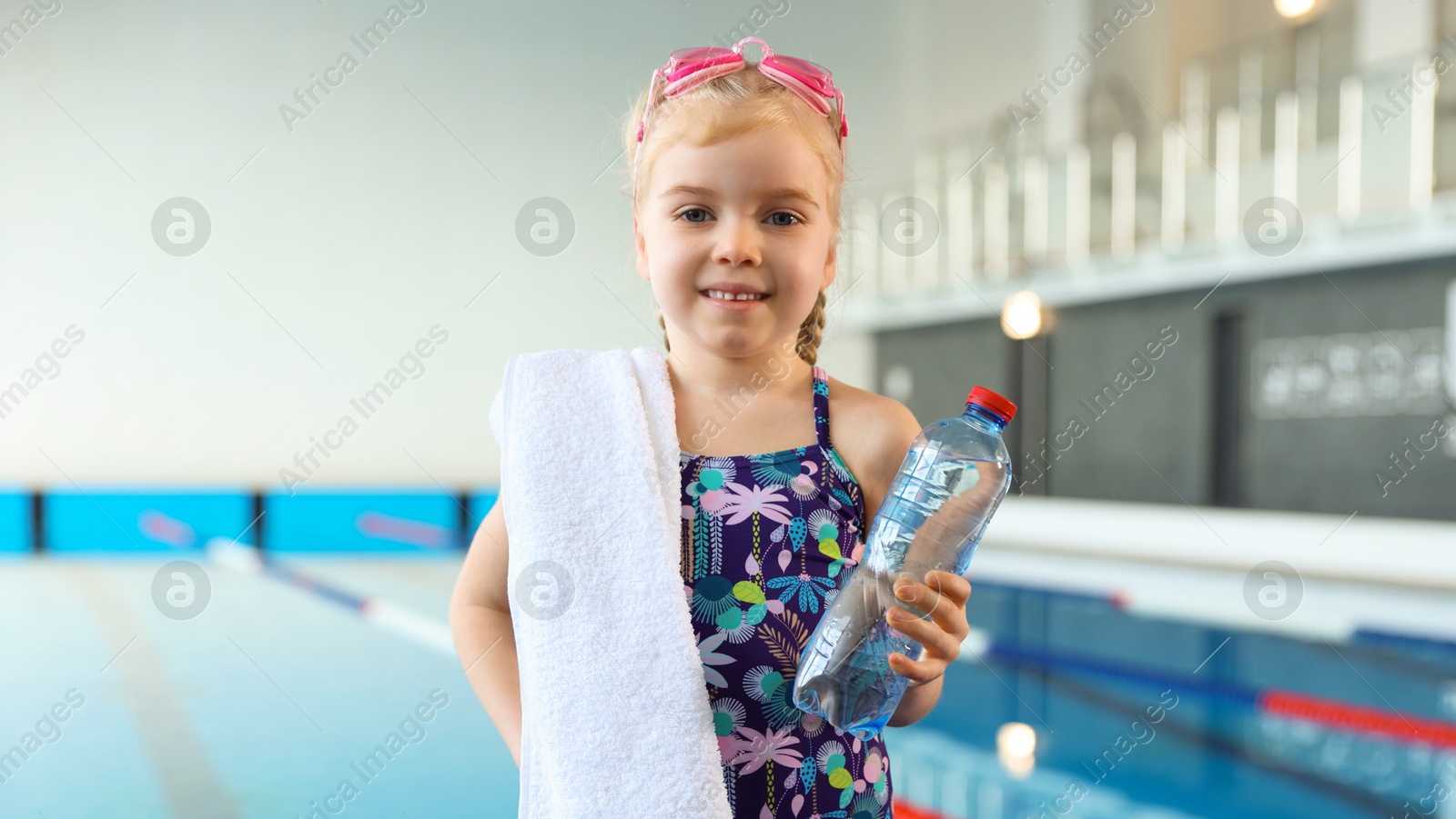 Photo of Little girl with goggles, towel and water near swimming pool indoors