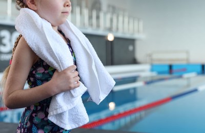 Photo of Little girl with goggles and towel near swimming pool indoors, closeup. Space for text