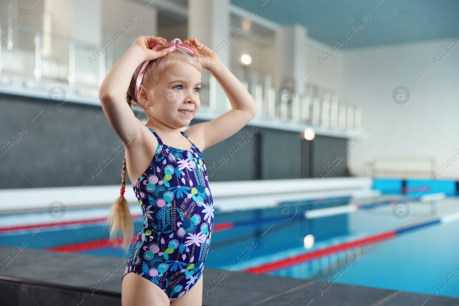 Photo of Little girl with goggles near swimming pool indoors, space for text