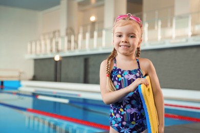 Photo of Little girl with goggles and kickboard near swimming pool indoors, space for text