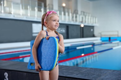 Photo of Little girl with goggles and kickboard near swimming pool indoors, space for text