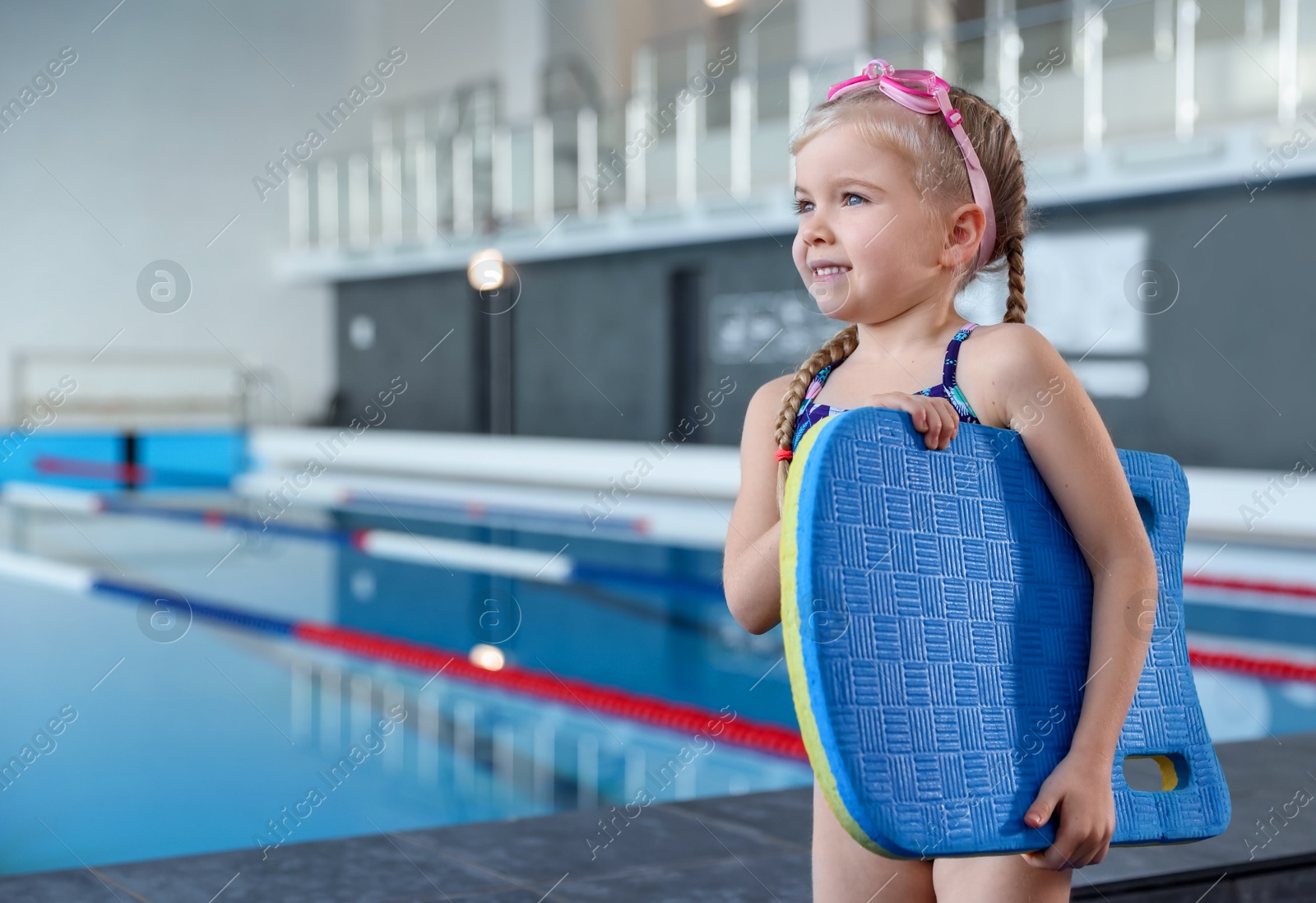 Photo of Little girl with goggles and kickboard near swimming pool indoors, space for text