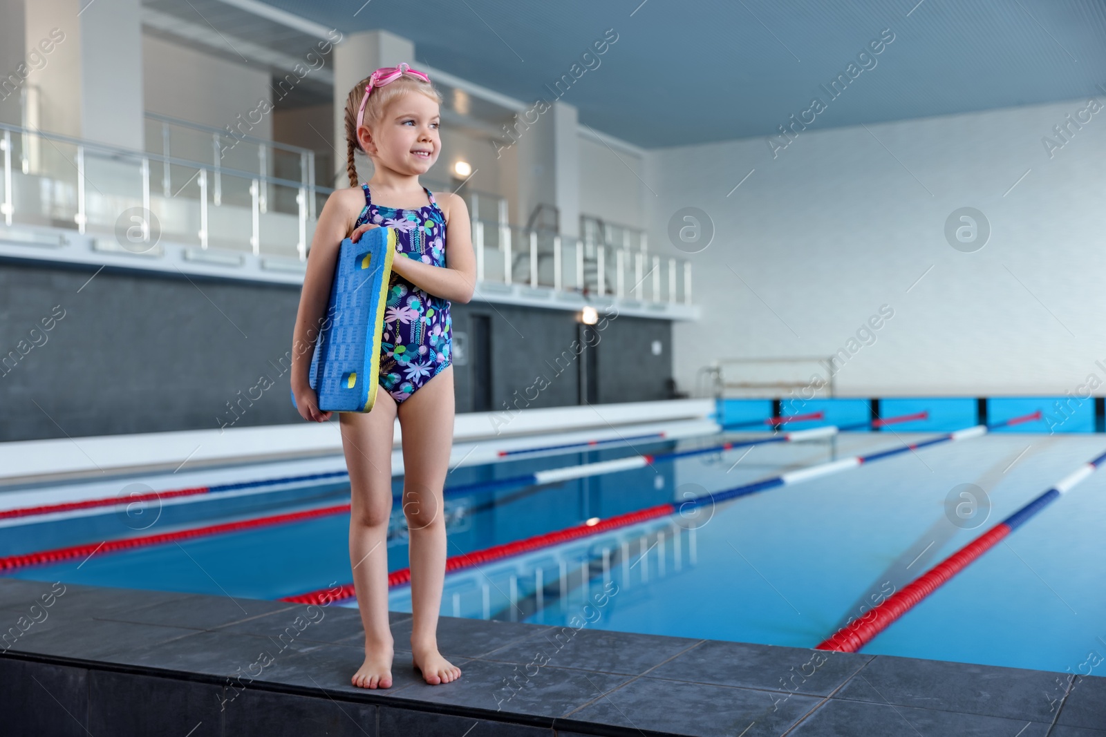 Photo of Little girl with goggles and kickboard near swimming pool indoors, space for text