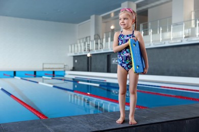 Photo of Little girl with goggles and kickboard near swimming pool indoors, space for text