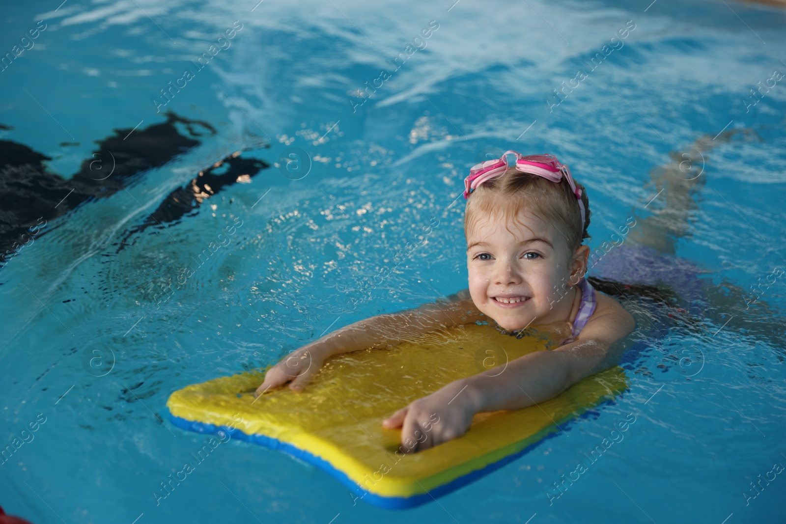 Photo of Little girl with goggles and kickboard swimming in pool indoors