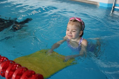 Photo of Little girl with goggles and kickboard swimming in pool indoors