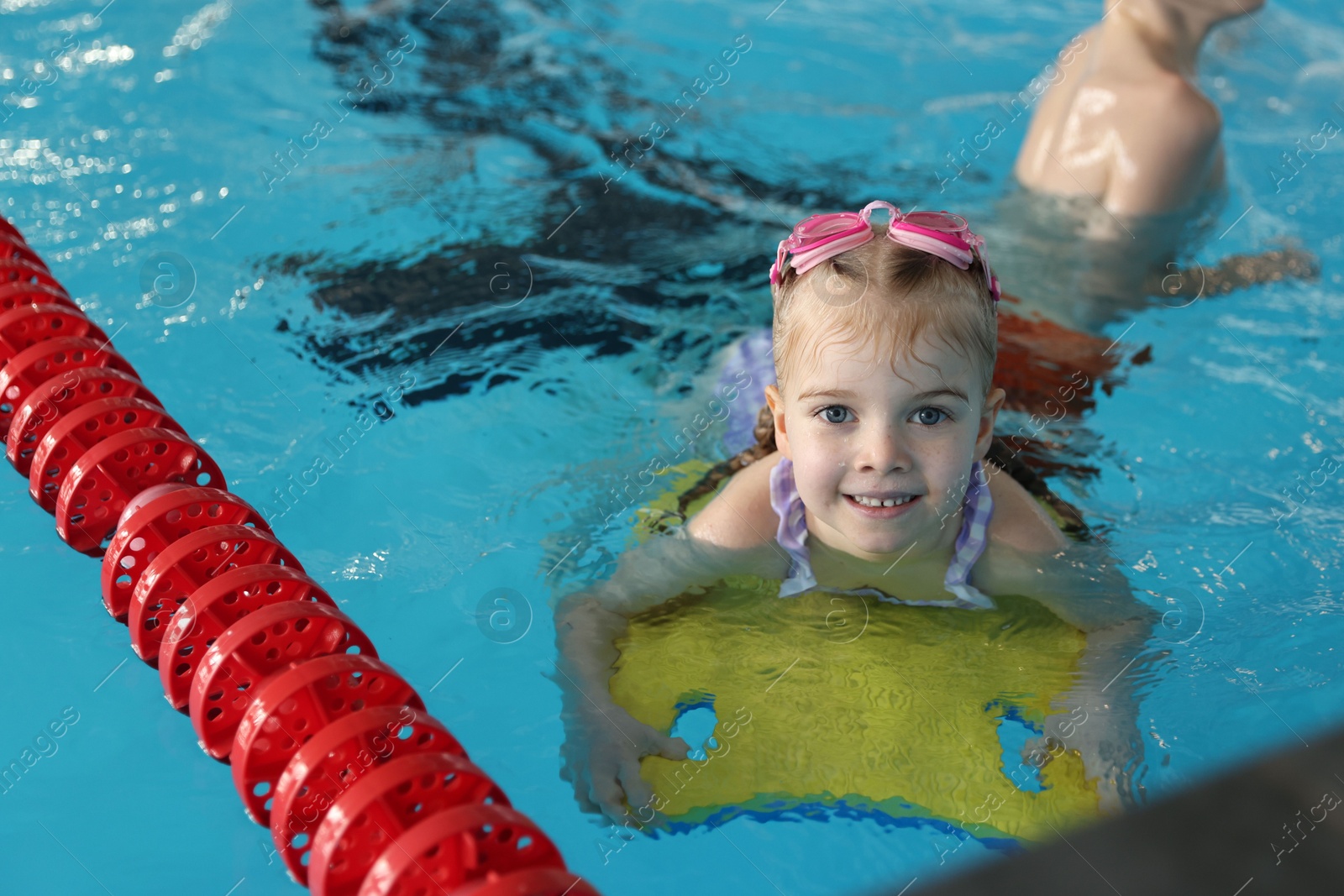 Photo of Little girl with goggles and kickboard swimming in pool indoors