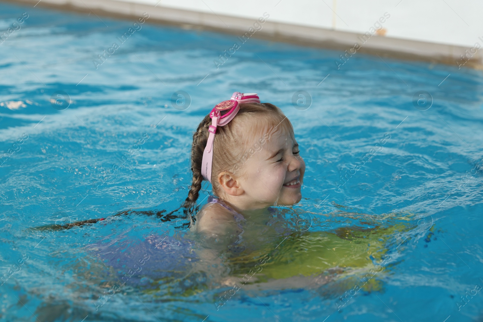 Photo of Little girl with goggles and kickboard swimming in pool indoors