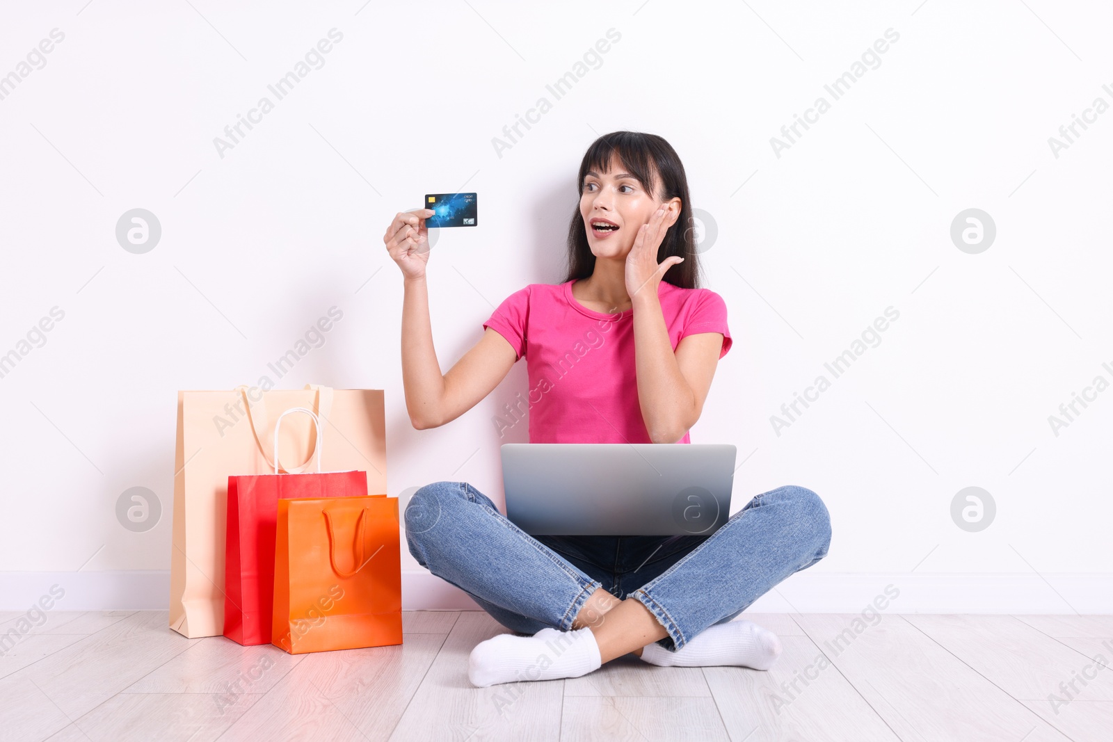 Photo of Internet shopping. Surprised woman with credit card, laptop and colorful bags sitting on floor near white wall