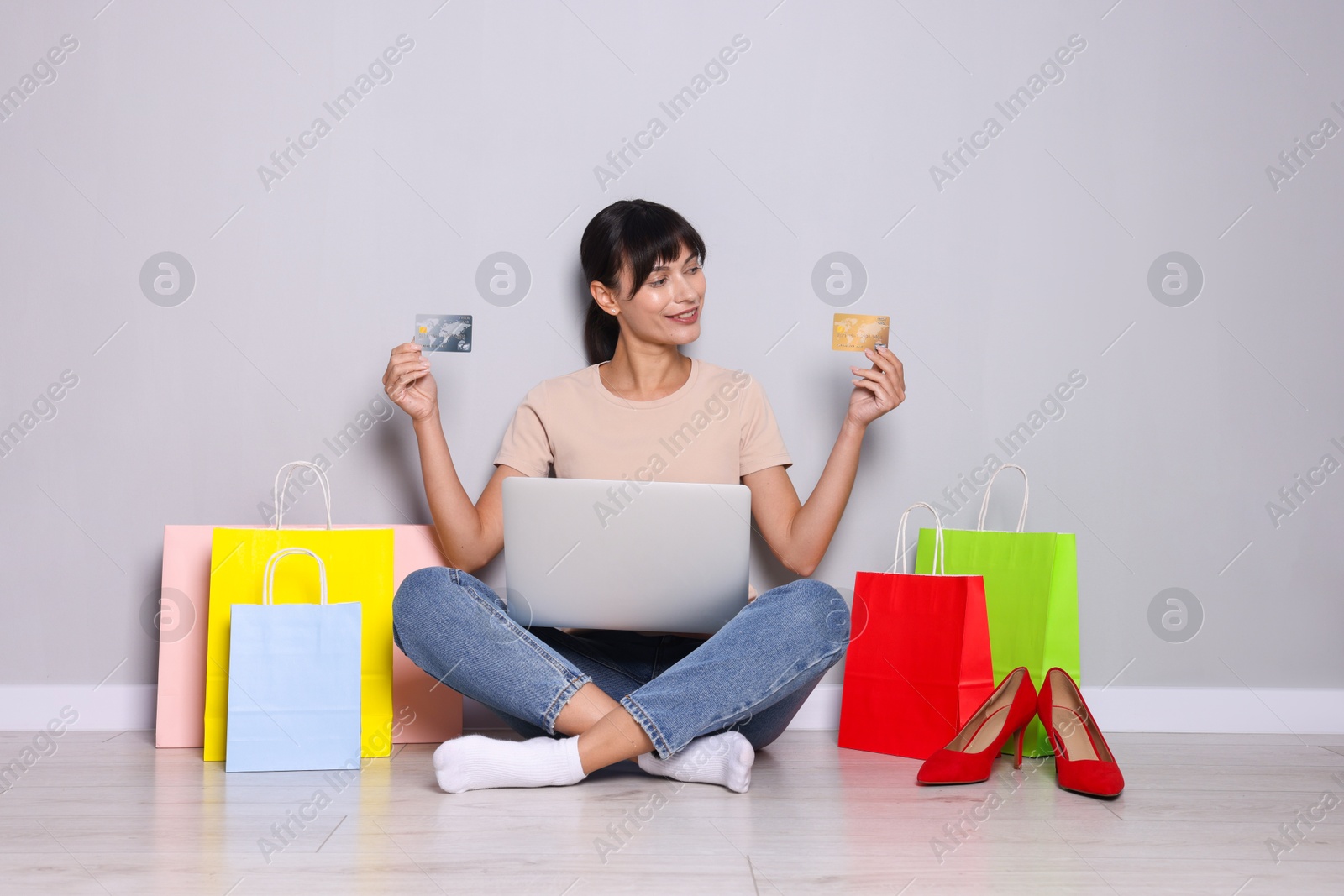 Photo of Internet shopping. Happy woman with credit cards, laptop and colorful bags sitting near grey wall