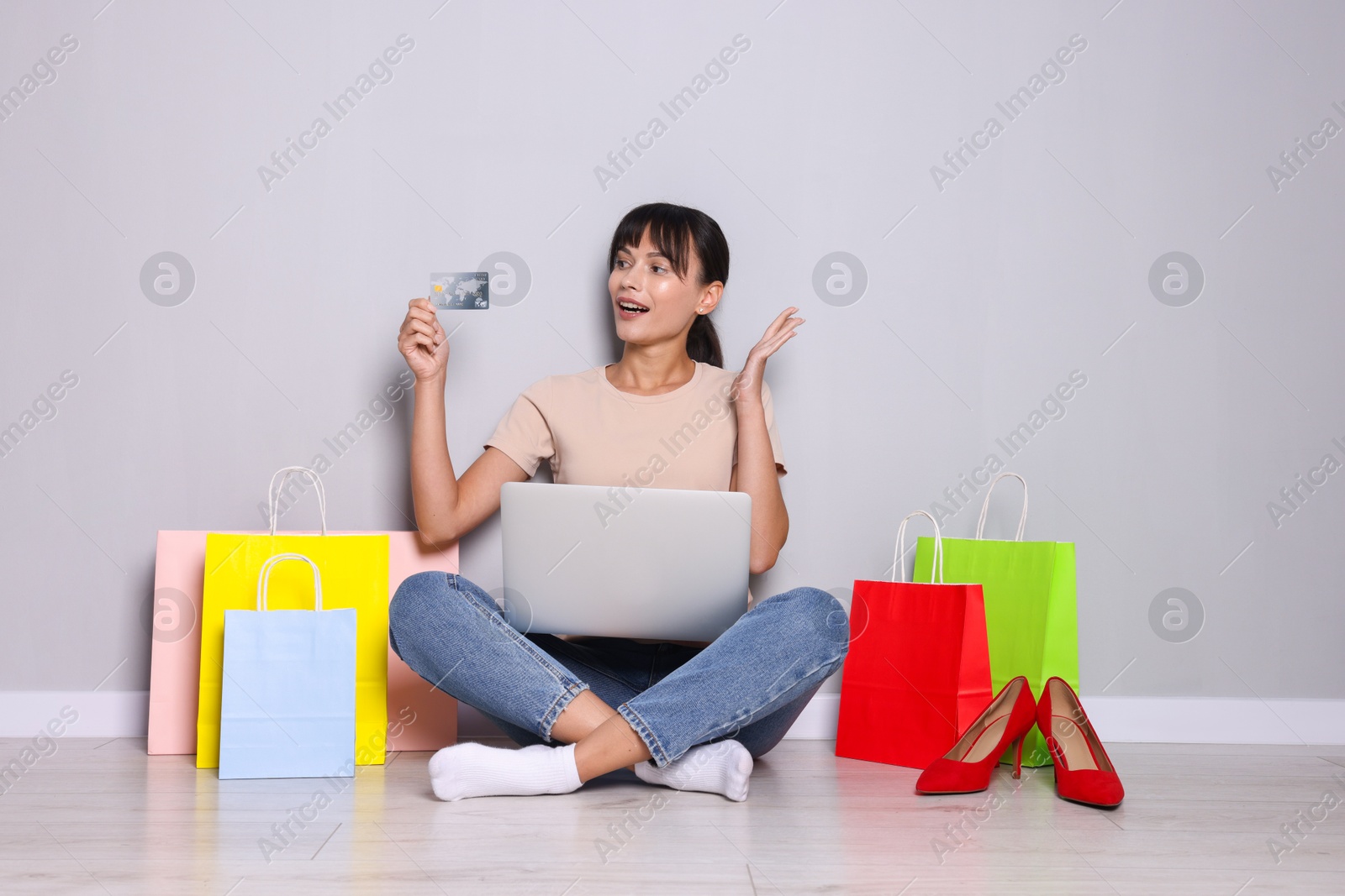 Photo of Internet shopping. Happy woman with credit card, laptop and colorful bags sitting near grey wall