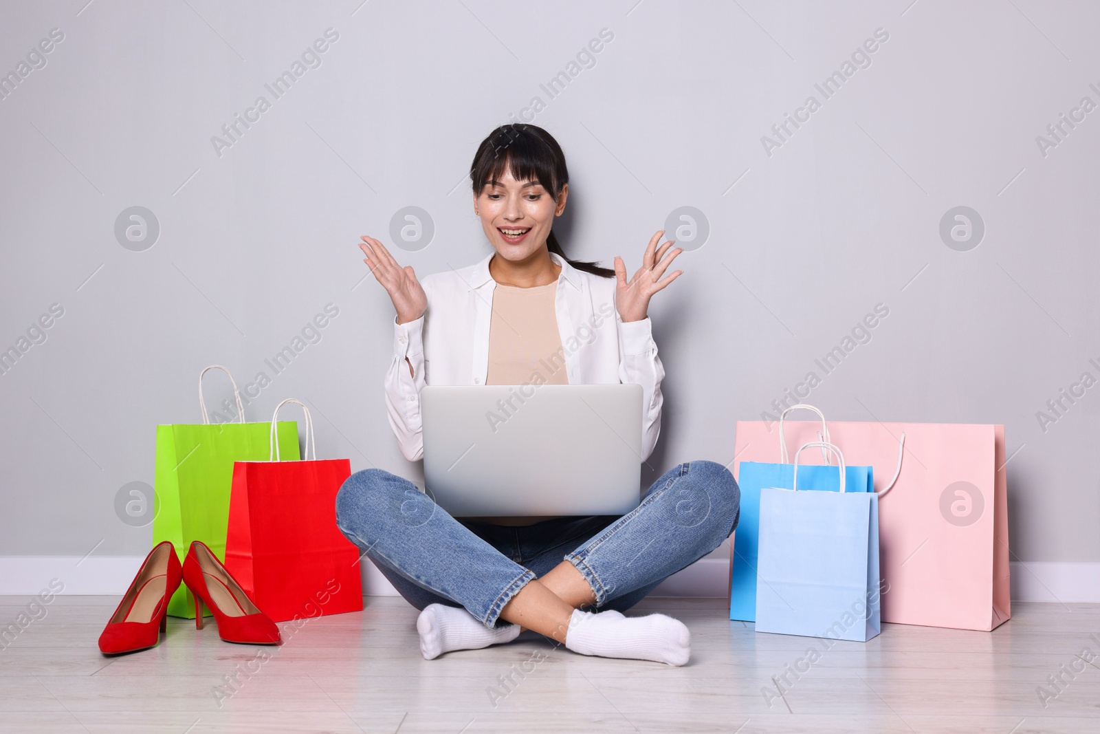 Photo of Internet shopping. Happy woman with laptop and colorful bags sitting near grey wall