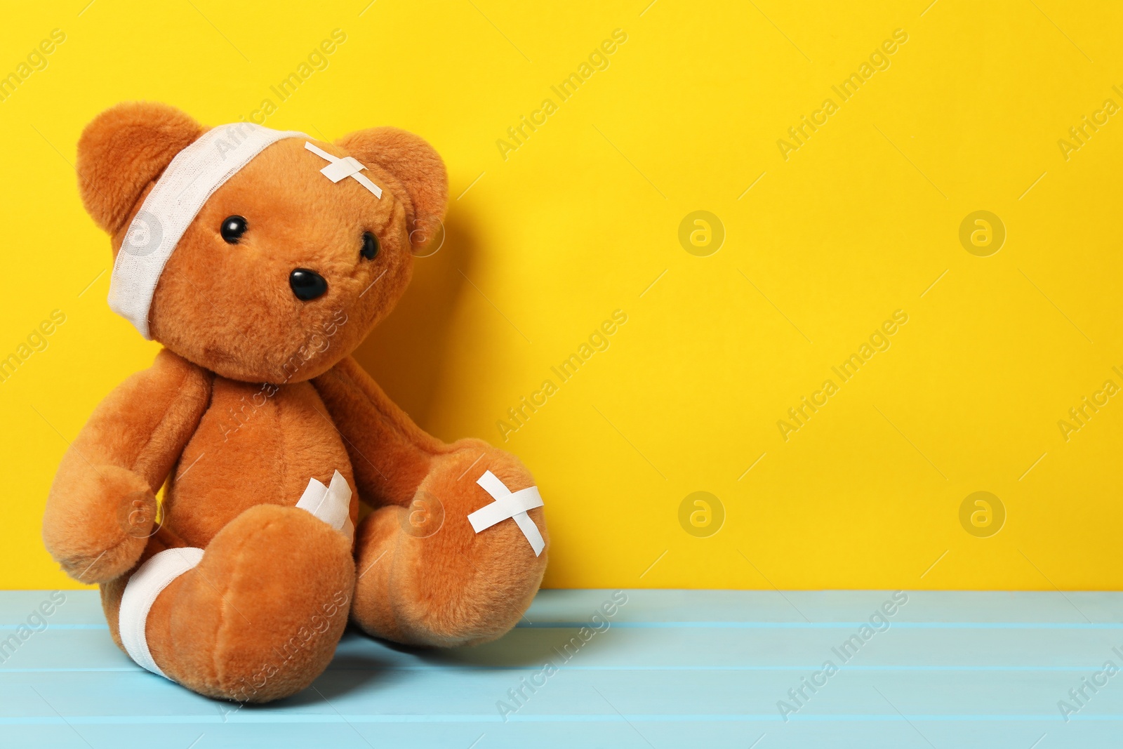 Photo of Teddy bear with bandages and sticking plasters on light blue wooden table against yellow background, space for text