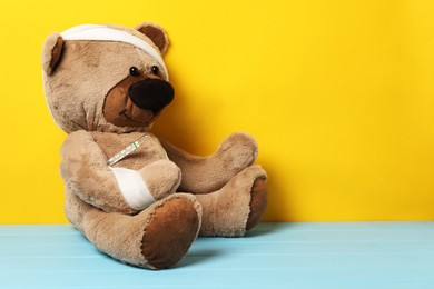 Photo of Teddy bear with bandages and thermometer on light blue wooden table against yellow background, space for text