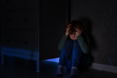 Scared boy hiding behind chest of drawers at night