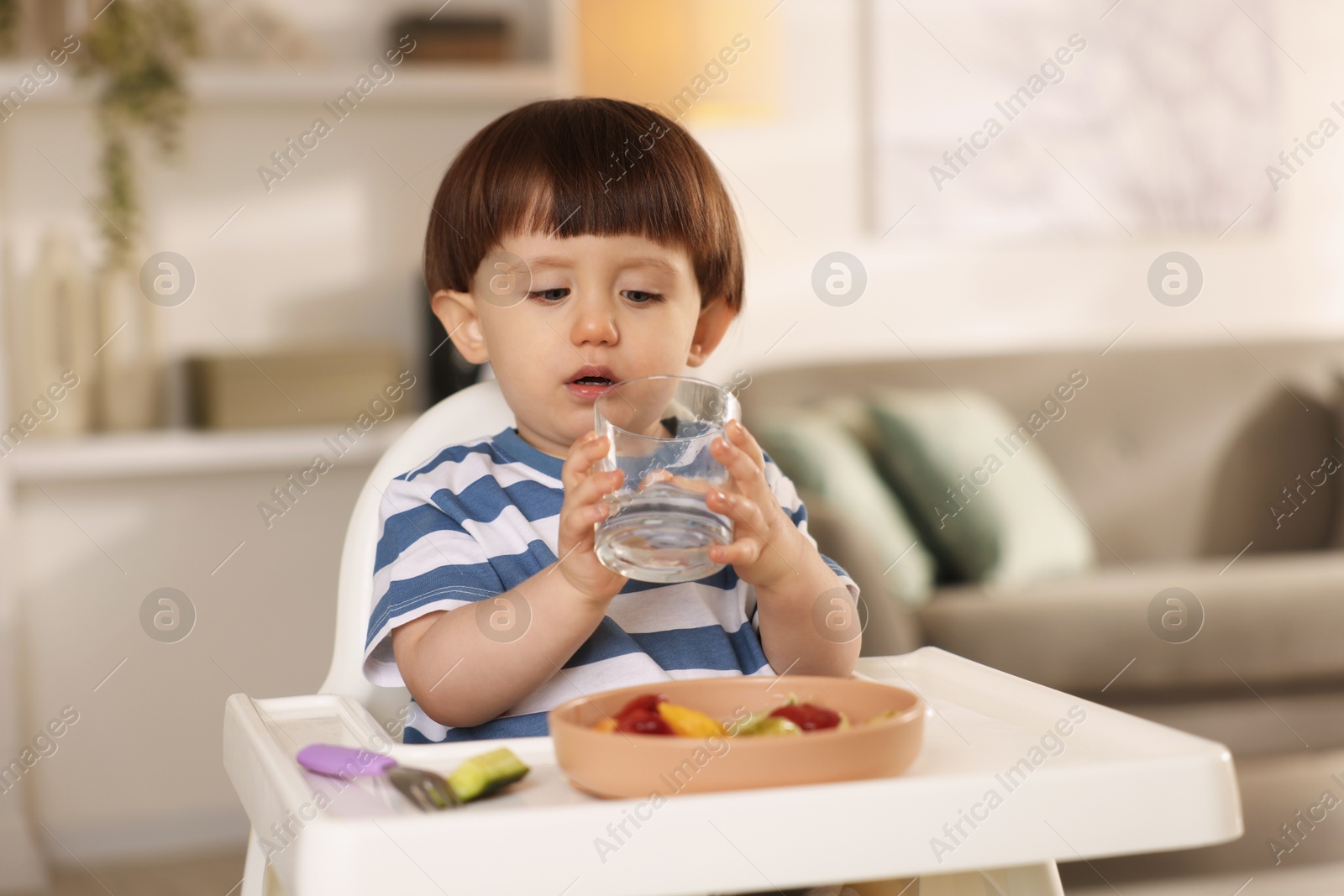 Photo of Cute little boy drinking water in high chair at home