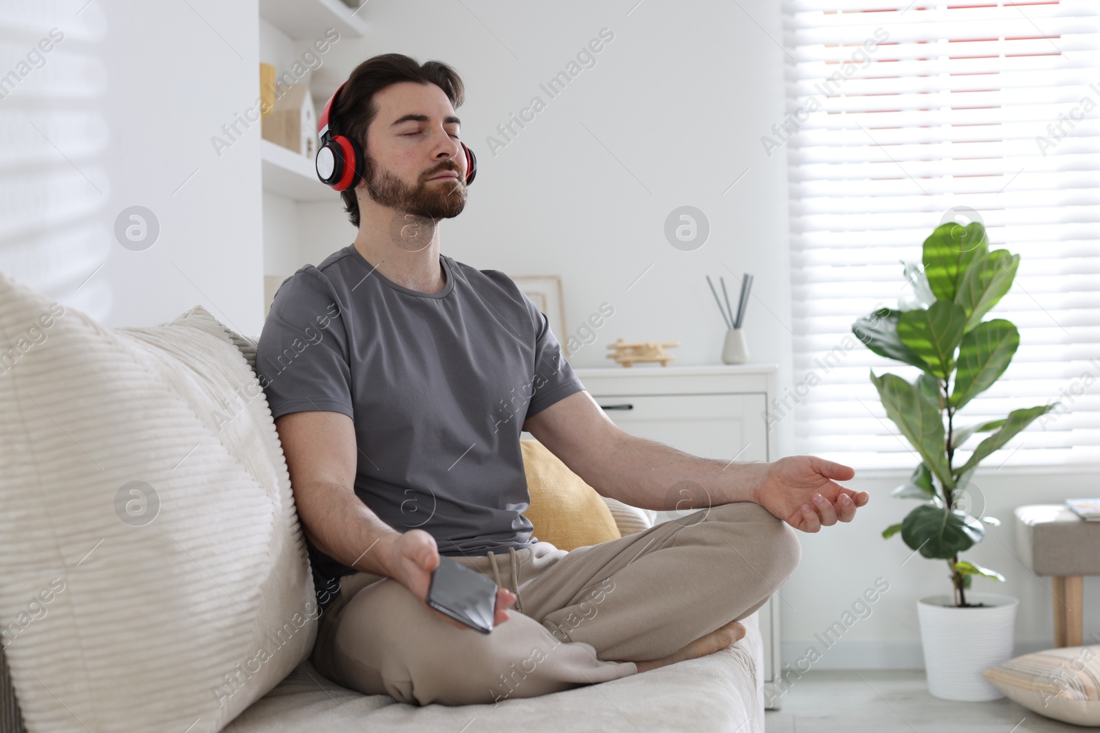 Photo of Handsome man in headphones with smartphone meditating on sofa at home