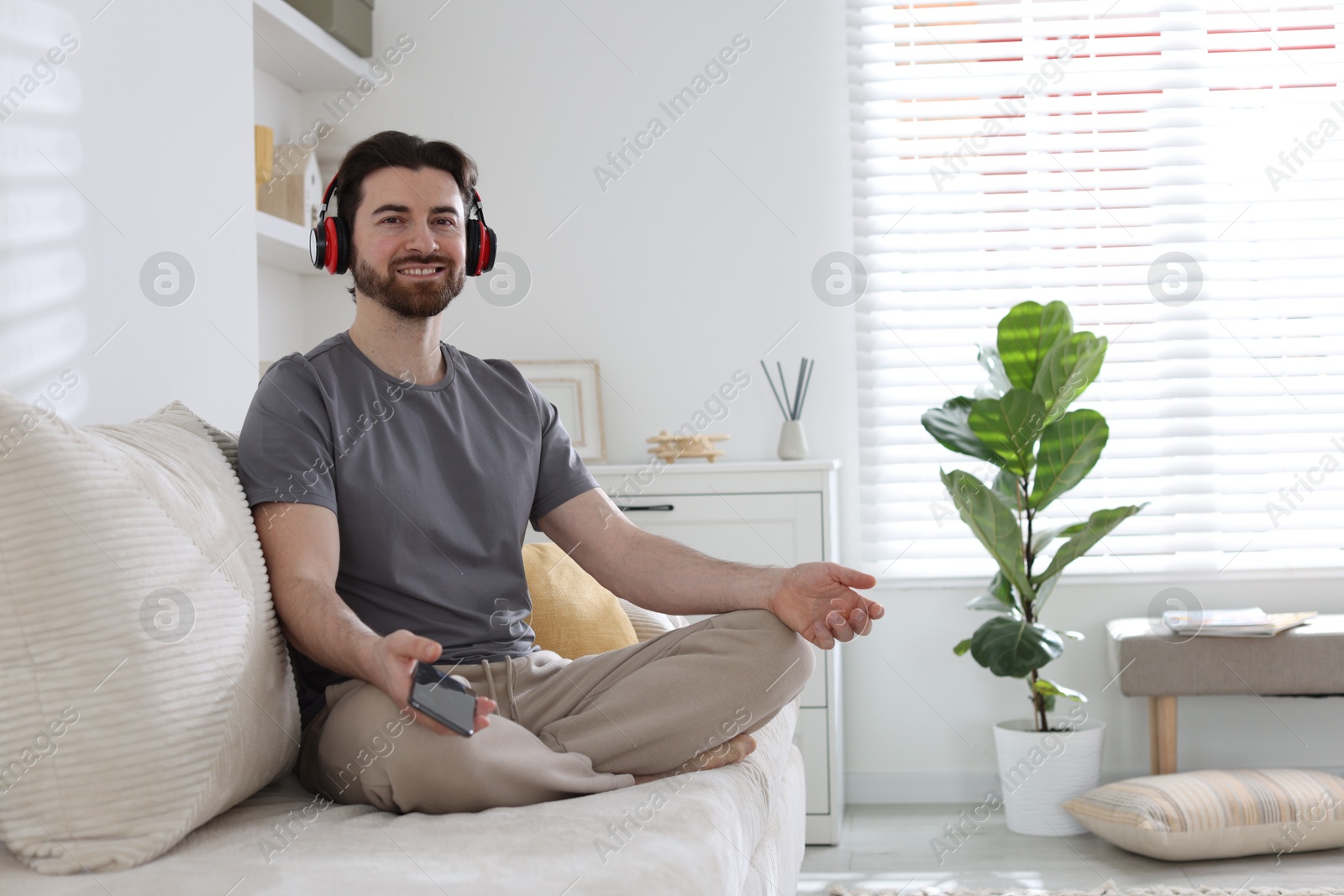 Photo of Smiling man in headphones with smartphone meditating on sofa at home