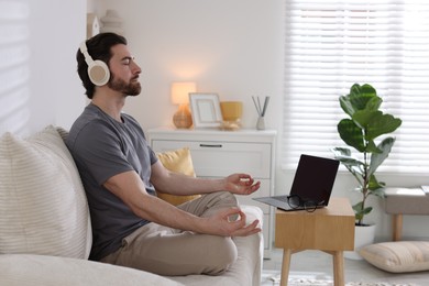 Photo of Man in headphones meditating near laptop on sofa at home