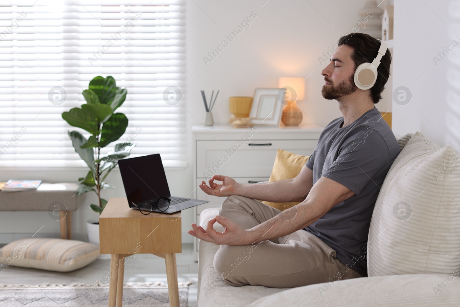 Photo of Man in headphones meditating near laptop on sofa at home