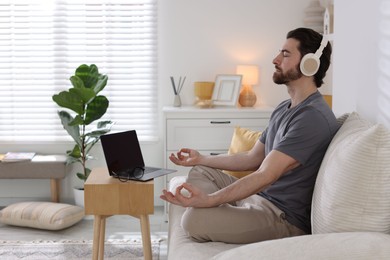 Photo of Man in headphones meditating near laptop on sofa at home