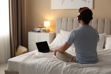 Photo of Man in headphones meditating near laptop on bed at home, back view