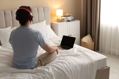 Photo of Man in headphones meditating near laptop on bed at home, back view