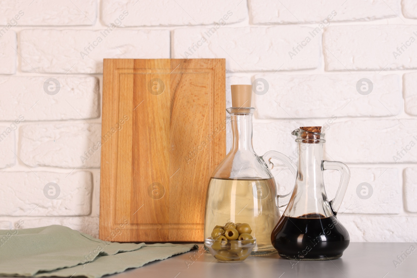 Photo of Bottles of salad dressings, olives and cutting board on table near white brick wall