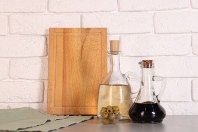 Photo of Bottles of salad dressings, olives and cutting board on table near white brick wall