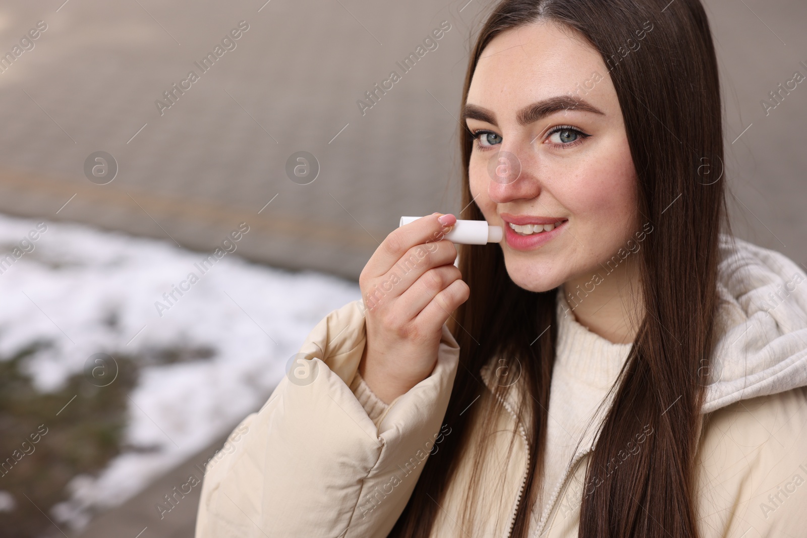 Photo of Beautiful young woman applying lip balm on winter day. Space for text