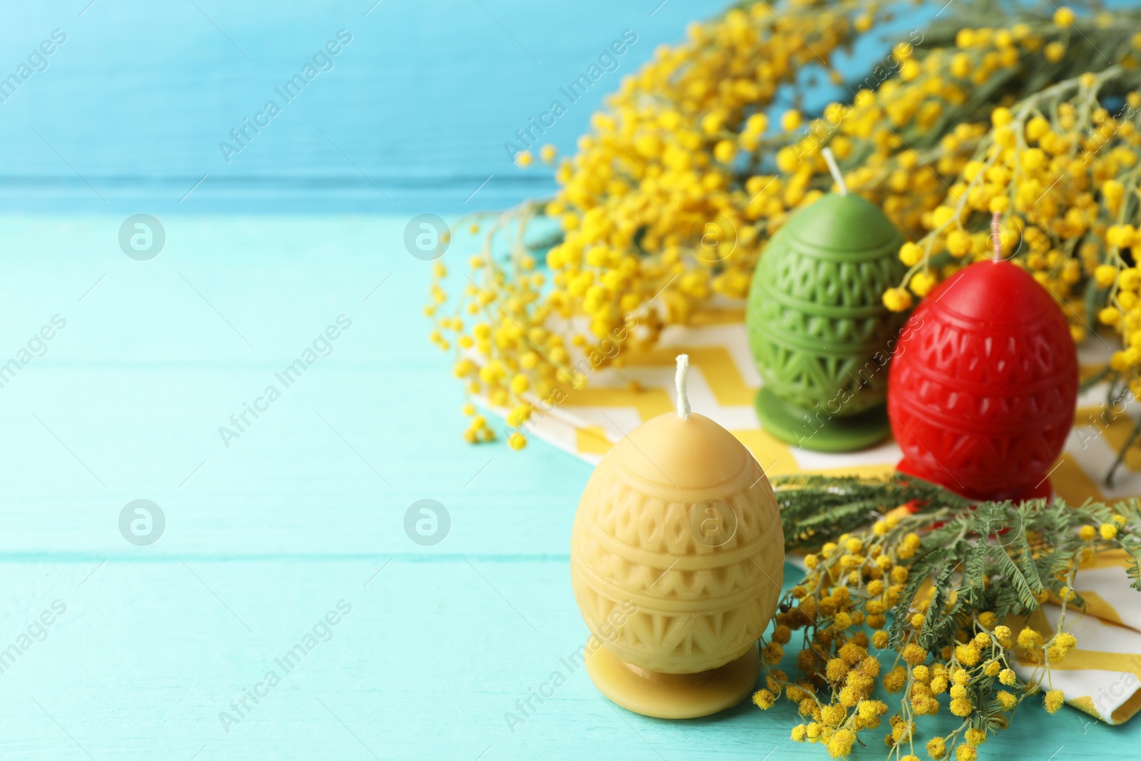 Photo of Colorful egg-shaped candles and mimosa flowers on turquoise wooden table against light blue background, closeup with space for text. Easter decor