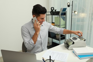 Photo of Man using banknote counter while talking on phone at white table indoors