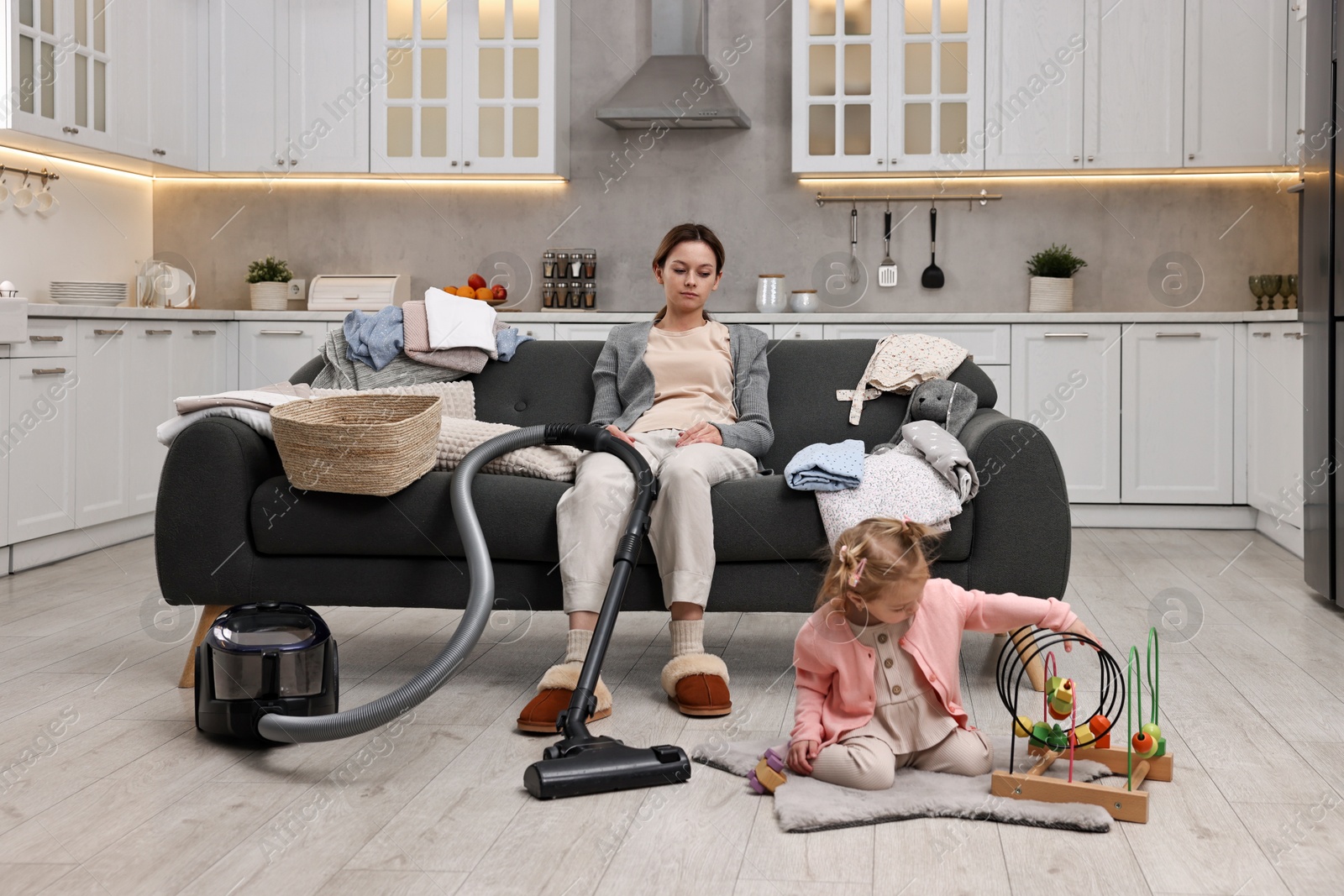 Photo of Tired housewife with vacuum cleaner sitting among messy laundry on sofa while her little daughter playing at home