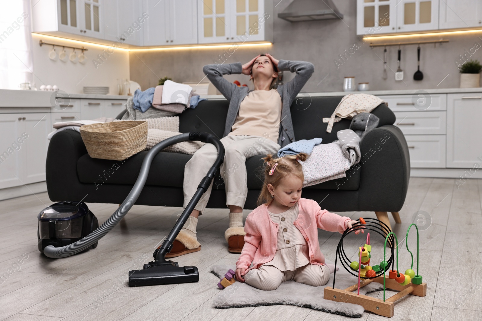 Photo of Tired housewife with vacuum cleaner sitting among messy laundry on sofa while her little daughter playing at home