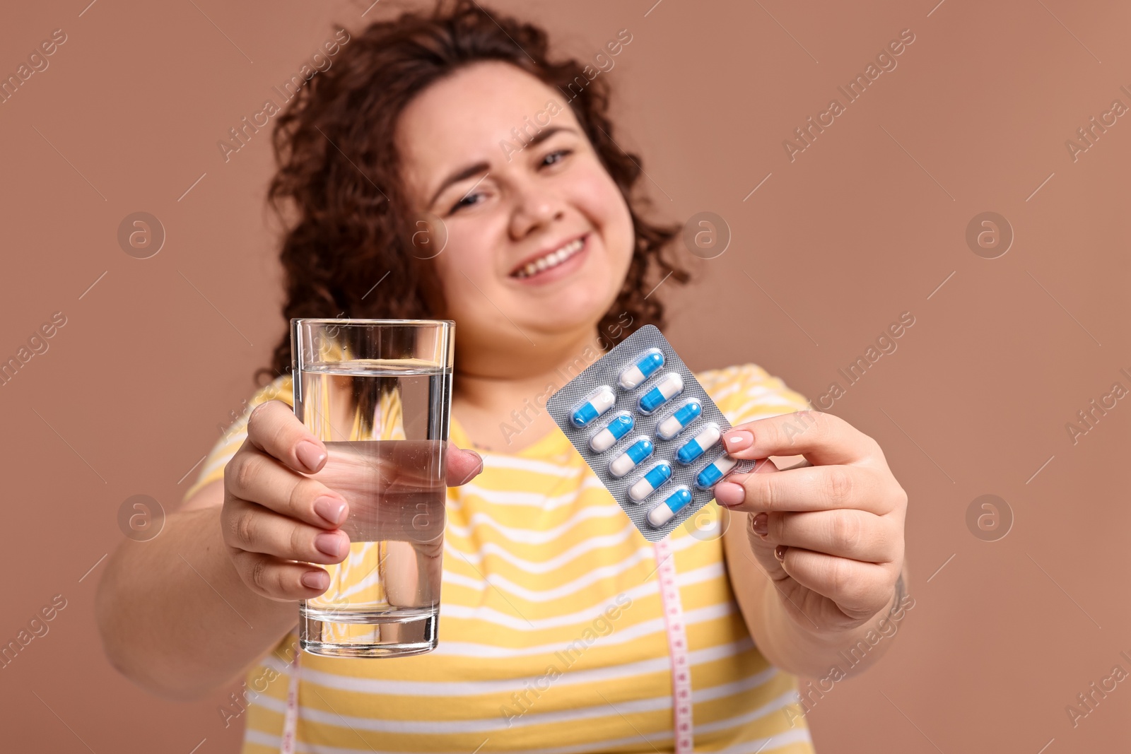 Photo of Happy plus size woman giving blister of weight loss supplements and glass of water on beige background, selective focus