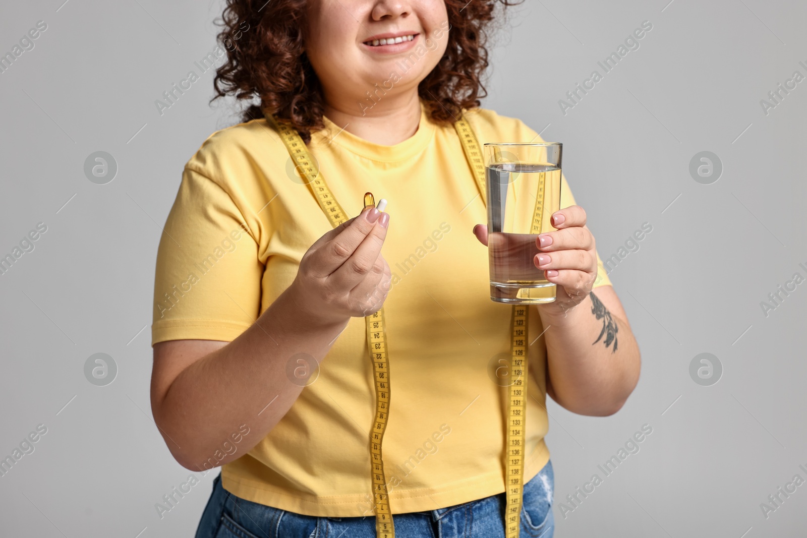 Photo of Happy plus size woman with weight loss supplements and glass of water on grey background, closeup