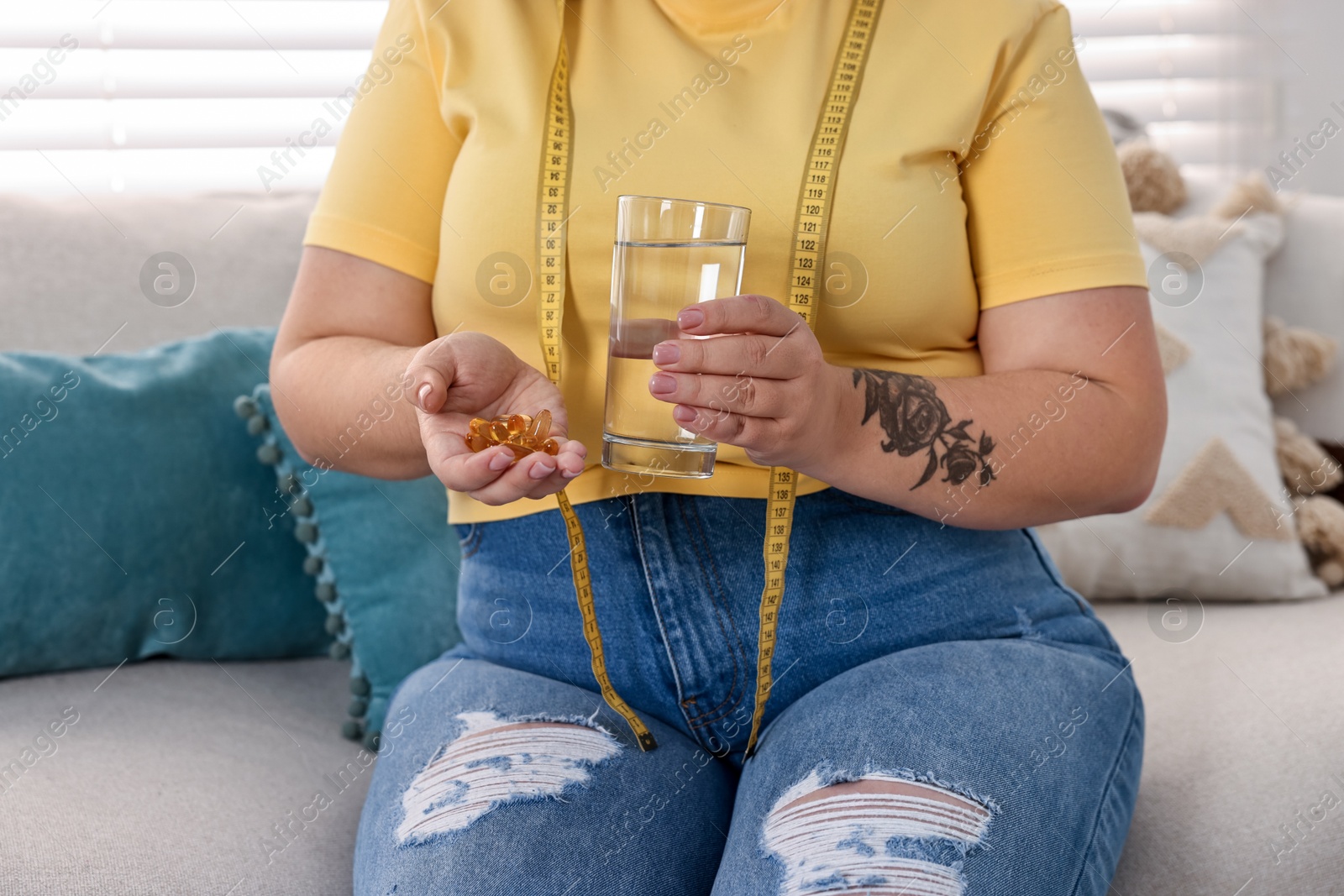 Photo of Plus size woman with pile of weight loss supplements and glass of water on sofa at home, closeup