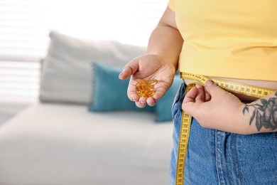 Photo of Plus size woman measuring waist with tape and holding pile of weight loss supplements at home, closeup. Space for text