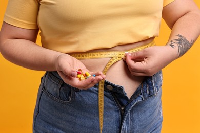 Photo of Plus size woman measuring waist with tape and holding pile of weight loss supplements on orange background, closeup