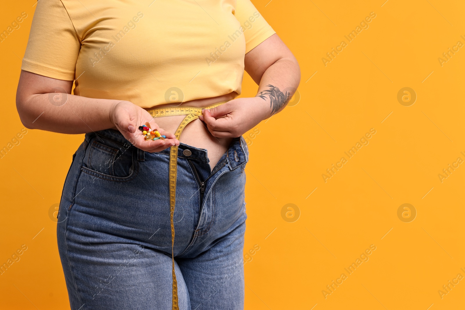 Photo of Plus size woman measuring waist with tape and holding pile of weight loss supplements on orange background, closeup. Space for text