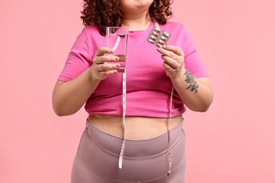 Photo of Plus size woman holding blister of weight loss supplements and glass of water on pink background, closeup