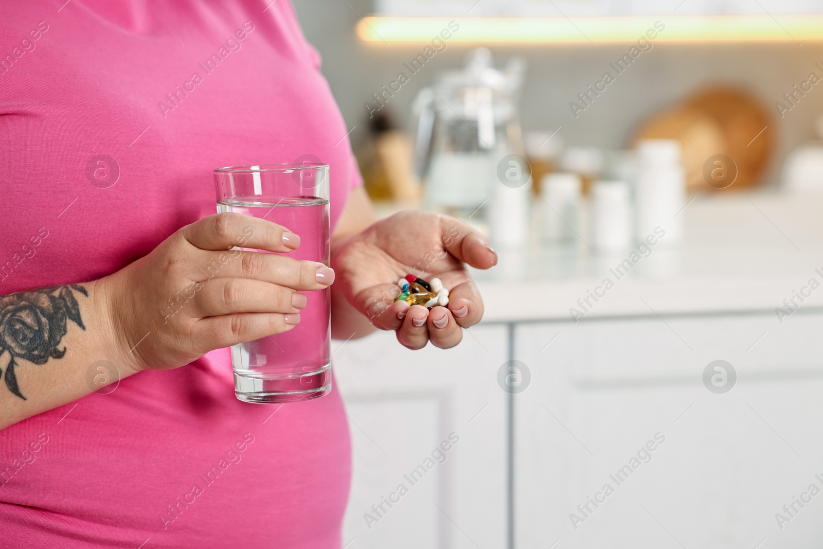 Photo of Plus size woman holding pile of weight loss supplements and glass with water in kitchen, closeup. Space for text