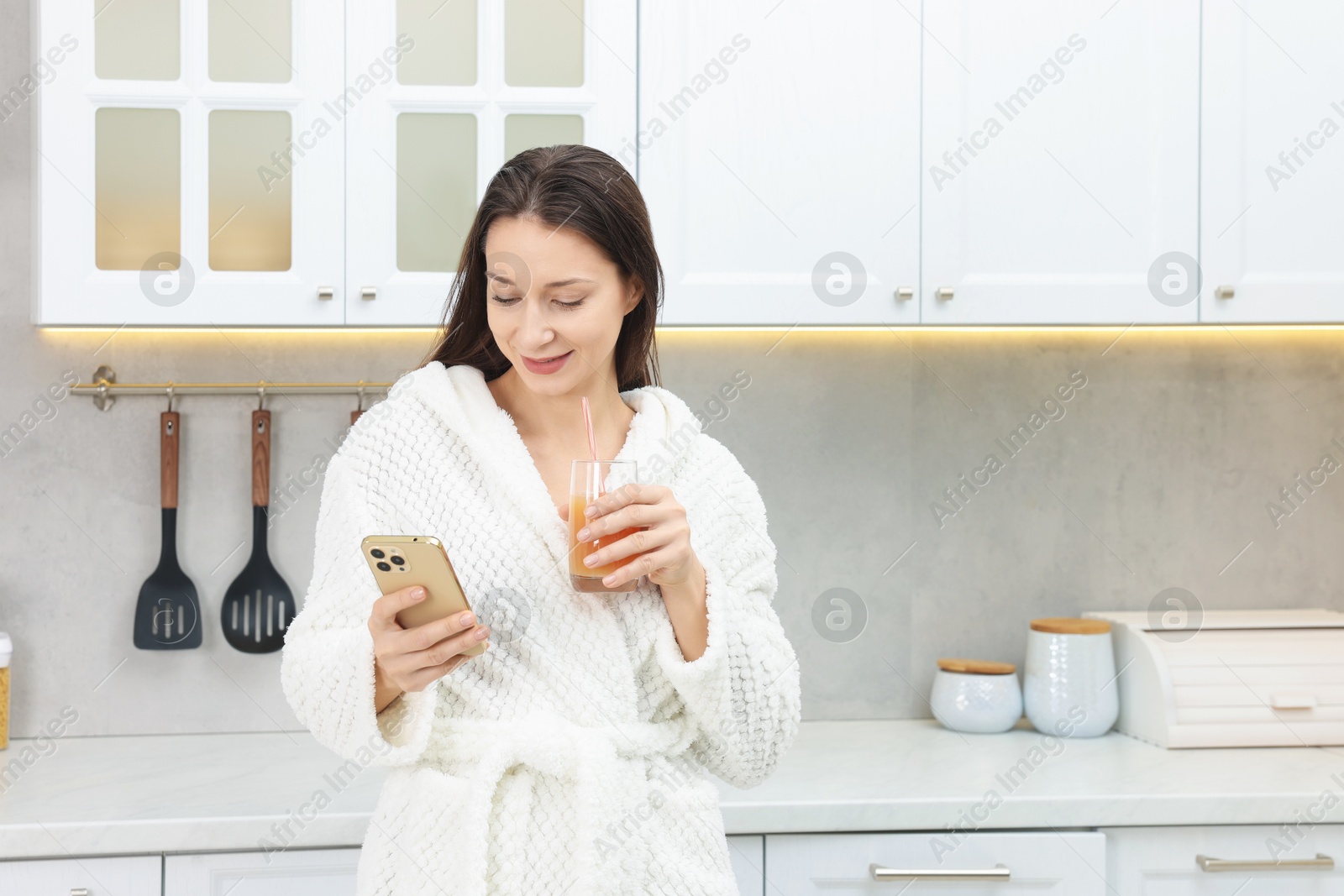 Photo of Beautiful woman with glass of juice using smartphone after spa procedure in kitchen