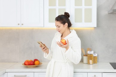 Photo of Beautiful woman with apple using smartphone after spa procedure in kitchen