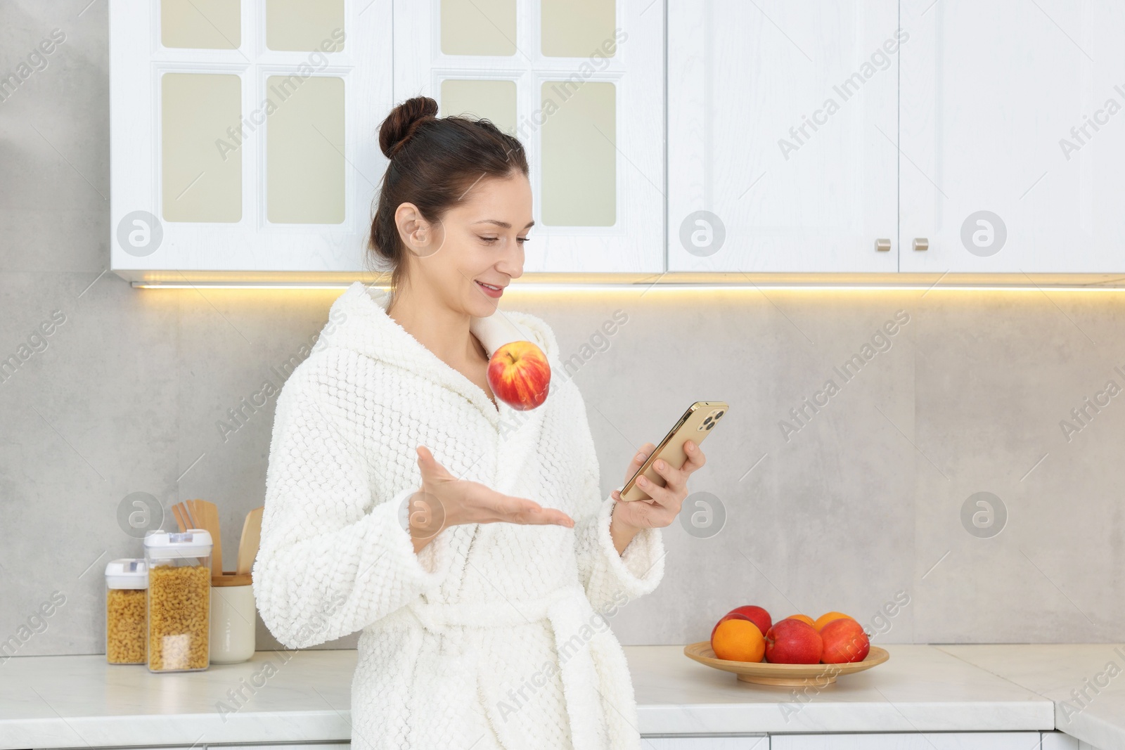 Photo of Beautiful woman throwing apple while using smartphone after spa procedure in kitchen