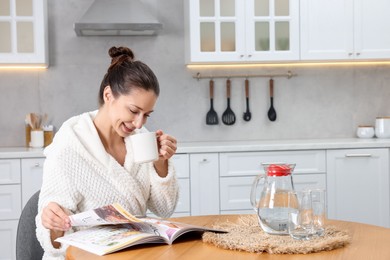 Photo of Beautiful woman with cup of drink reading magazine after spa procedure at wooden table in kitchen