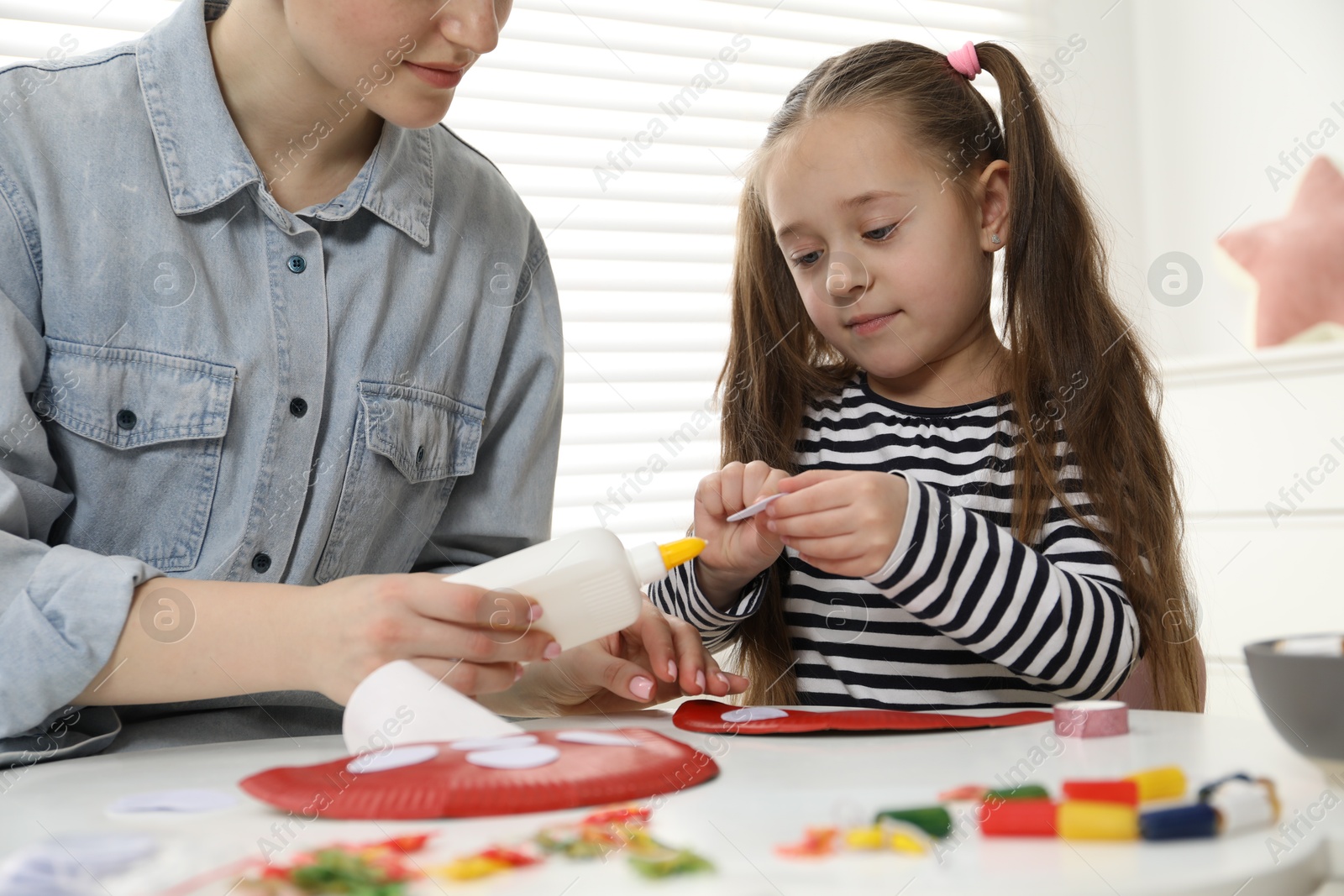 Photo of Woman and little girl making paper fly agaric at white table indoors, closeup. Child handmade craft