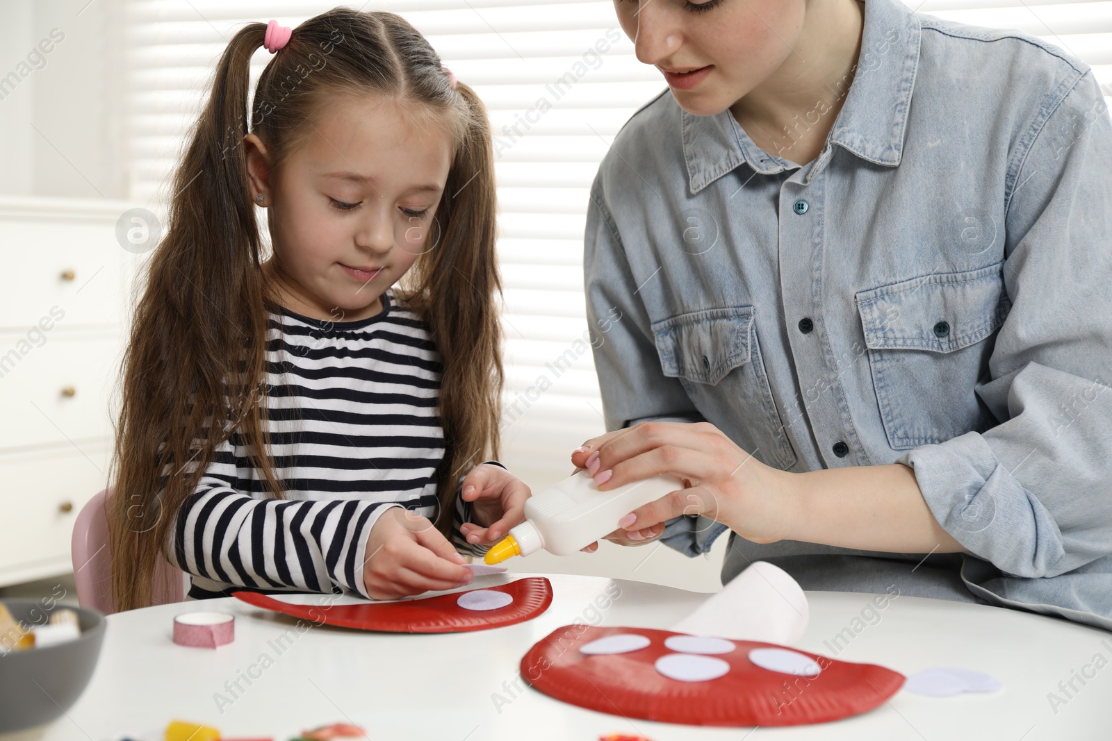 Photo of Woman and little girl making paper fly agaric at white table indoors. Child handmade craft