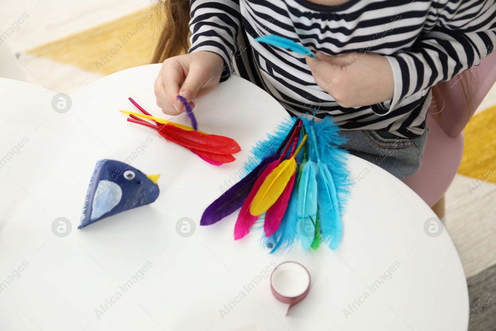 Photo of Little girl making paper bird at white table indoors, closeup. Child handmade craft