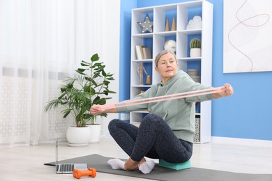 Photo of Elderly woman exercising with fitness elastic band near laptop at home